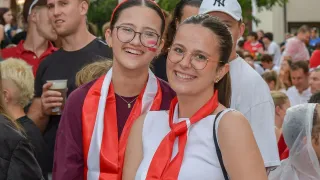 Zwei Frauen in roten und weißen Schärpen und rotem Gesichtsmake-up stehen lächelnd zusammen. Eine trägt eine Brille und eine rote Krawatte, während die andere österreichische Flaggen in den Haaren hat. Hinter ihnen sind Menschen verschwommen.