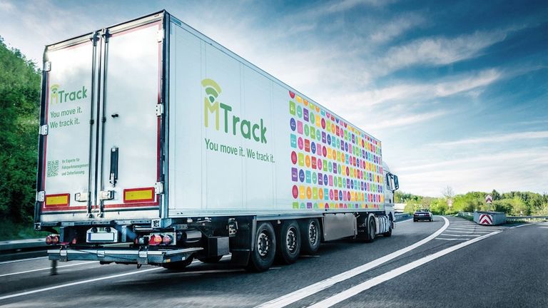 Ein großer LKW zeigt das MTTrack-Logo an seiner Seite. Es steht You move it. We track it. Der LKW ist auf einer Autobahn mit einem Auto dahinter.