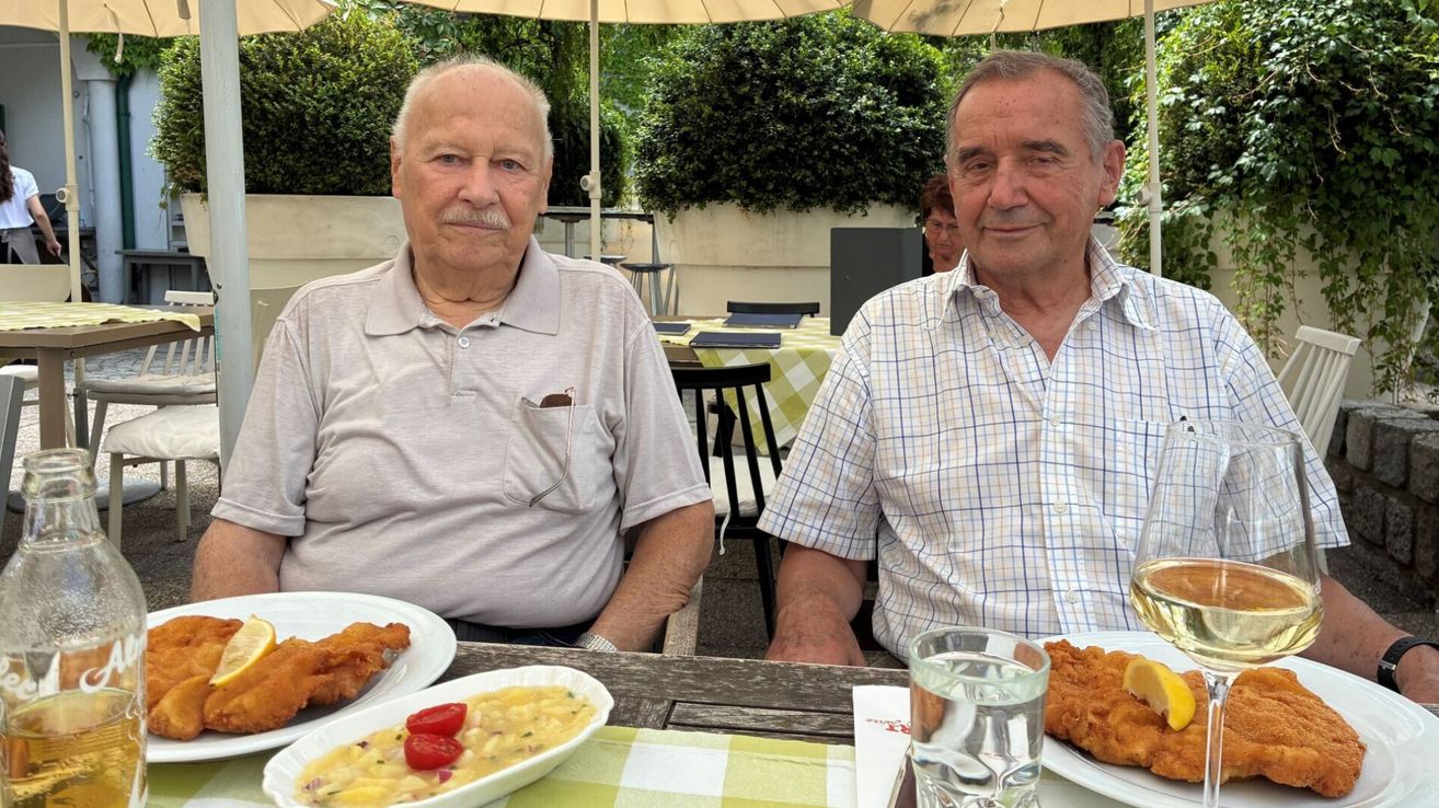 Zwei Männer sitzen an einem Tisch draußen. Einer hat einen Teller mit Essen und ein Glas Wasser, der andere ein Glas Wasser. Sie lächeln.