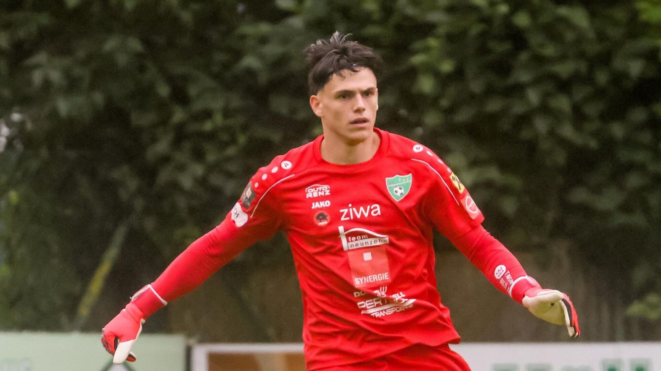 Ein Fußballspieler in einem roten Trikot mit 'ziwa' und 'SYNnergie'-Aufnähern läuft mit Bäumen im Hintergrund vorwärts.