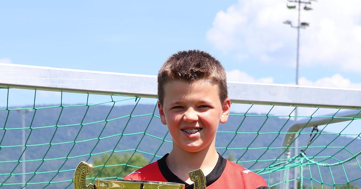 Ein Junge mit Zahnspange hält einen goldenen Pokal vor einem Fußballtor. Er lächelt in die Kamera.