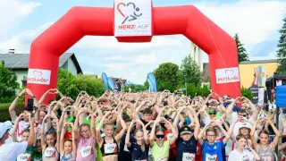 Die Teilnehmer eines Marathons stehen unter einem roten Torbogen mit Herzsymbol und bilden Herzformen mit ihren Händen. Bäume und Häuser sind im Hintergrund zu sehen.