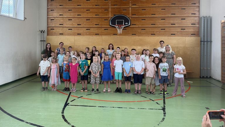 Eine Gruppe von Kindern und Erwachsenen steht in einer Turnhalle und posiert für ein Foto. Ein Basketballkorb ist an der Holzwand hinter ihnen montiert.