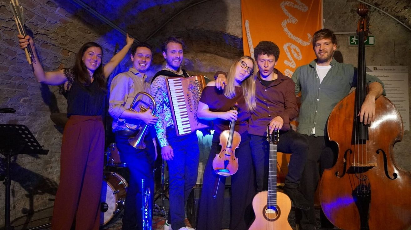 Eine Gruppe von Musikern posiert für ein Foto und hält verschiedene Instrumente. Dazu gehören eine Trompete, ein Akkordeon, eine Geige und eine Gitarre. Der Hintergrund zeigt ein orangefarbenes Banner mit weißen Schriftzügen.