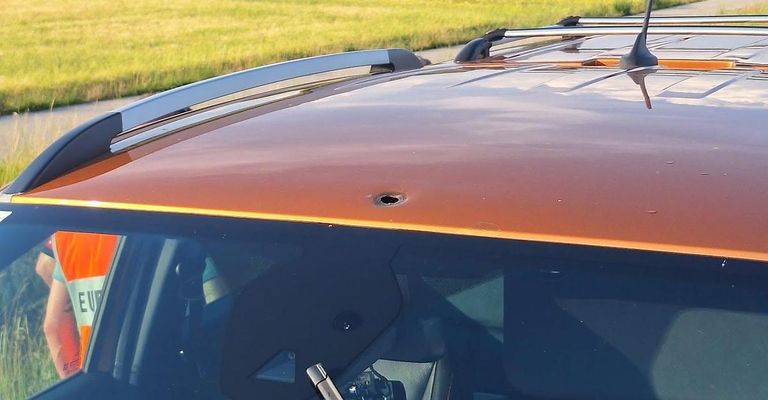 Das orangefarbene Dach eines Autos zeigt ein Loch von einer Kugel. Der Dachträger ist silbern. Drinnen ist die Windschutzscheibe klar. Das Autoinnere ist dunkel.
