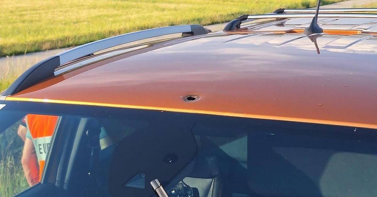 Das orangefarbene Dach eines Autos zeigt ein Loch von einer Kugel. Der Dachträger ist silbern. Drinnen ist die Windschutzscheibe klar. Das Autoinnere ist dunkel.
