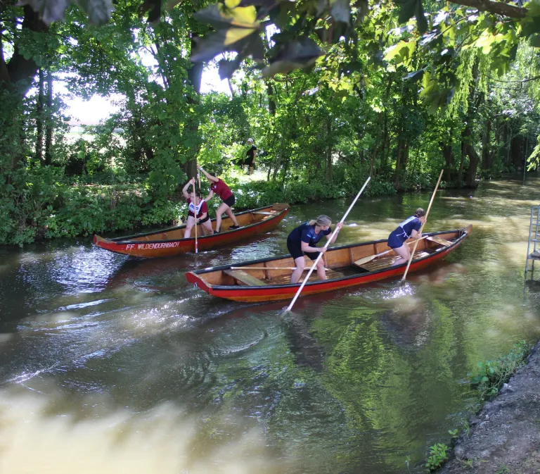 Zwei Personen paddeln mit Holzbooten auf einem Fluss. Die Boote haben rote Besatze und eines hat Text an der Seite. Bäume umgeben den Fluss.