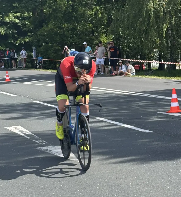 Ein Radfahrer in einem roten Trikot fährt während eines Rennens auf der Straße mit einem Fahrrad, wobei Zuschauer zusehen und orangefarbene Kegel an der Seite stehen.