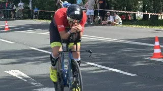 Ein Radfahrer in einem roten Trikot fährt während eines Rennens auf der Straße mit einem Fahrrad, wobei Zuschauer zusehen und orangefarbene Kegel an der Seite stehen.