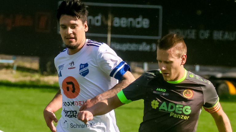 Zwei Fußballspieler auf einem Feld. Einer ist in Weiß und der andere in Schwarz, beide in intensivem Spiel. Der Spieler in Weiß hat ein Logo mit Text, und der Spieler in Schwarz hat das Logo eines Sponsors.