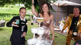 Ein Mann und eine Frau gießen Champagner in eine Pyramide von Gläsern bei einer Veranstaltung im Freien. Die Frau lächelt, und es gibt Menschen, Bäume und einen Schirm im Hintergrund.