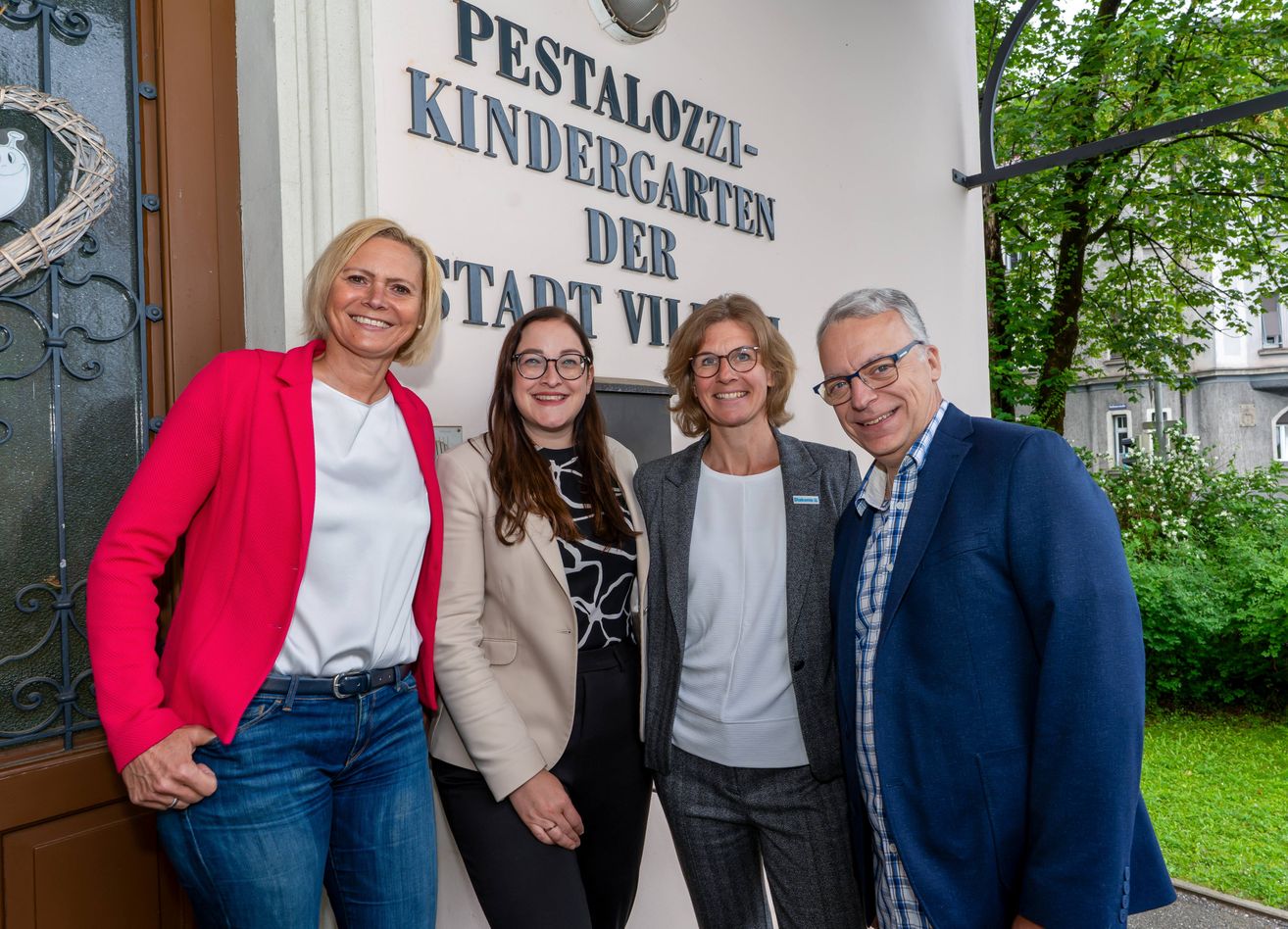 Fünf Personen in formeller Kleidung stehen lächelnd vor einem Gebäude für ein Foto. Das Schild sagt Pestalozzi Kindergarten der Stadt Vill. Bäume und eine Lampe sind im Hintergrund sichtbar.