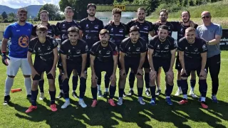 Eine Rugbymannschaft in schwarzen Trikots mit verschiedenfarbigen Socken posiert für ein Foto auf einem grünen Feld, mit einem Schild im Hintergrund, das 'GAST' steht.
