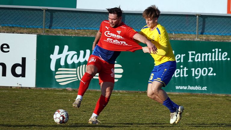 Zwei Fußballspieler sind in einem Spiel auf einem Feld. Einer trägt ein rotes Trikot und der andere gelb. Sie kämpfen um den Ball.