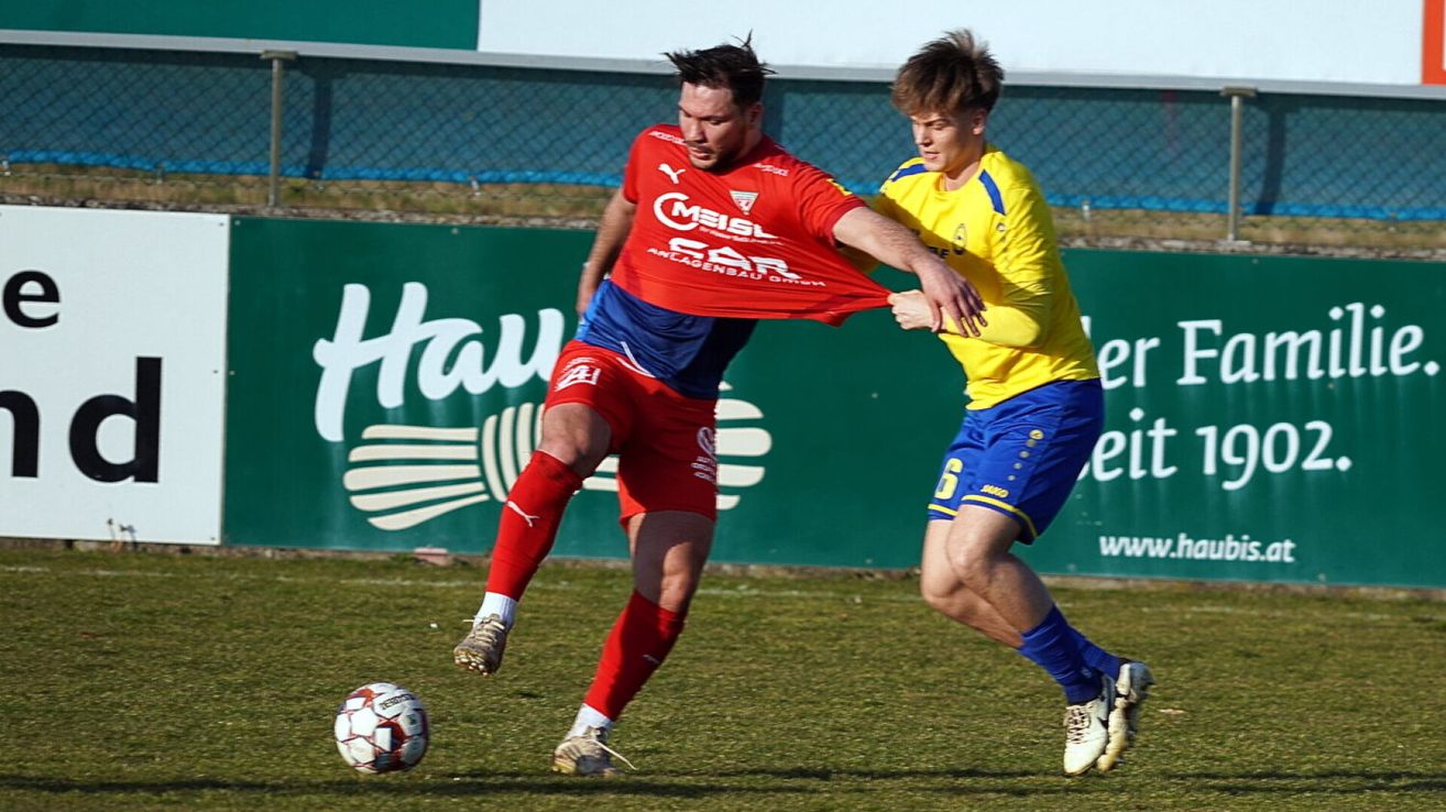 Zwei Fußballspieler sind in einem Spiel auf einem Feld. Einer trägt ein rotes Trikot und der andere gelb. Sie kämpfen um den Ball.