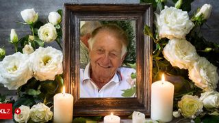 Ein Foto eines älteren Mannes mit einem lächelnden Gesicht, das in einem Spiegel reflektiert wird, umgeben von weißen Blumen und brennenden Kerzen.