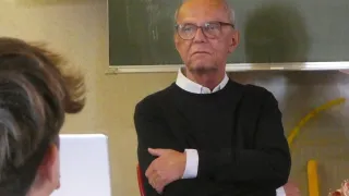 Ein älterer Mann mit Brille sitzt im Klassenzimmer, die Arme verschränkt, ernst blickend.