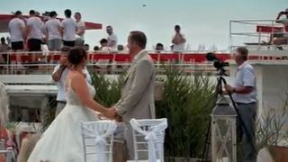 Eine Hochzeitszeremonie findet im Freien statt. Das Paar hält Händchen. Dahinter sind Gäste und ein Videograf. Die Zeremonie ist von Grünpflanzen umgeben.