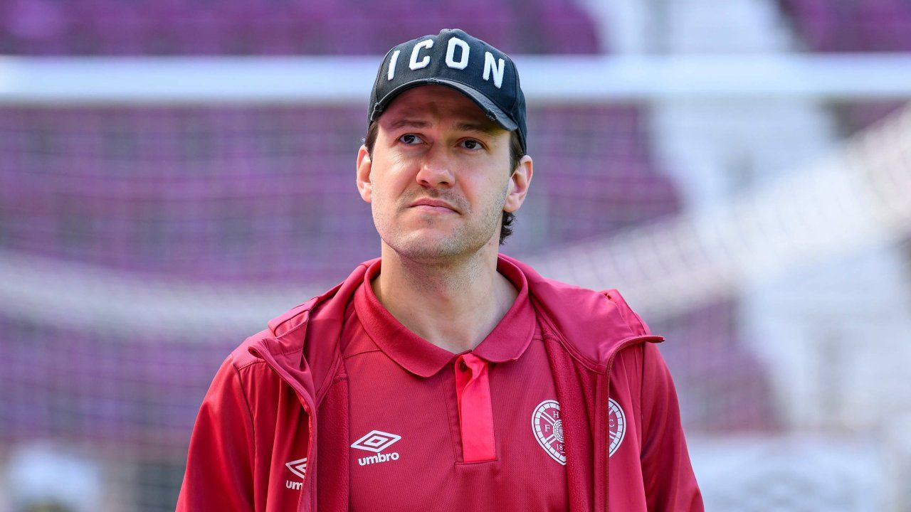 Ein Mann in einem roten Jackett mit dem Wort ICON auf einem schwarzen Baseball-Cap schaut direkt in die Kamera. Der Hintergrund ist unscharf mit einem Zaun und lila Sitzen.