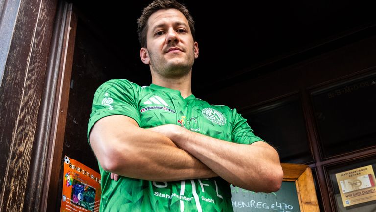Ein Mann mit ernstem Gesichtsausdruck trägt ein grünes Fußballtrikot mit Sponsorenlogos an den Armen.