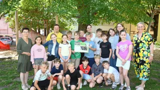 Eine Gruppe von Kindern und Erwachsenen steht in einem Park unter einem Baum. Einige Kinder halten ein Plakat. Dahinter befindet sich ein Gebäude mit einer Treppe.