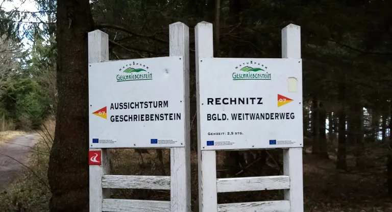 Zwei weiße Schilder auf Pfosten in einem Wald. Ein Schild steht 'AUSSICHTSTORM GESCHRIEBENSTEIN'. Das andere Schild zeigt 'RECHNITZ BGL. WEITANDERWEG'. Beide befinden sich im Naturpark Geschriebestein.
