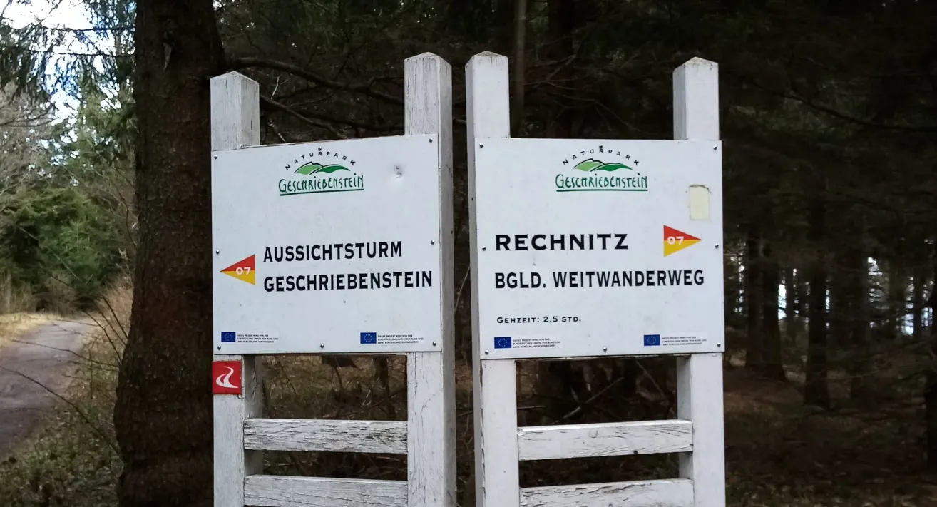 Zwei weiße Schilder auf Pfosten in einem Wald. Ein Schild steht 'AUSSICHTSTORM GESCHRIEBENSTEIN'. Das andere Schild zeigt 'RECHNITZ BGL. WEITANDERWEG'. Beide befinden sich im Naturpark Geschriebestein.