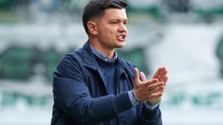 Ein Mann in einer blauen Jacke macht Handbewegungen auf einem Feld, wahrscheinlich ein Trainer, der Anweisungen gibt.