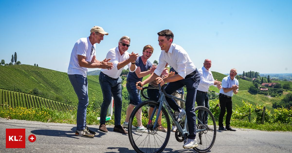 Eine Gruppe von Männern steht um einen Radfahrer auf einer Straße in einem Weinberg und lächelt und klatscht.