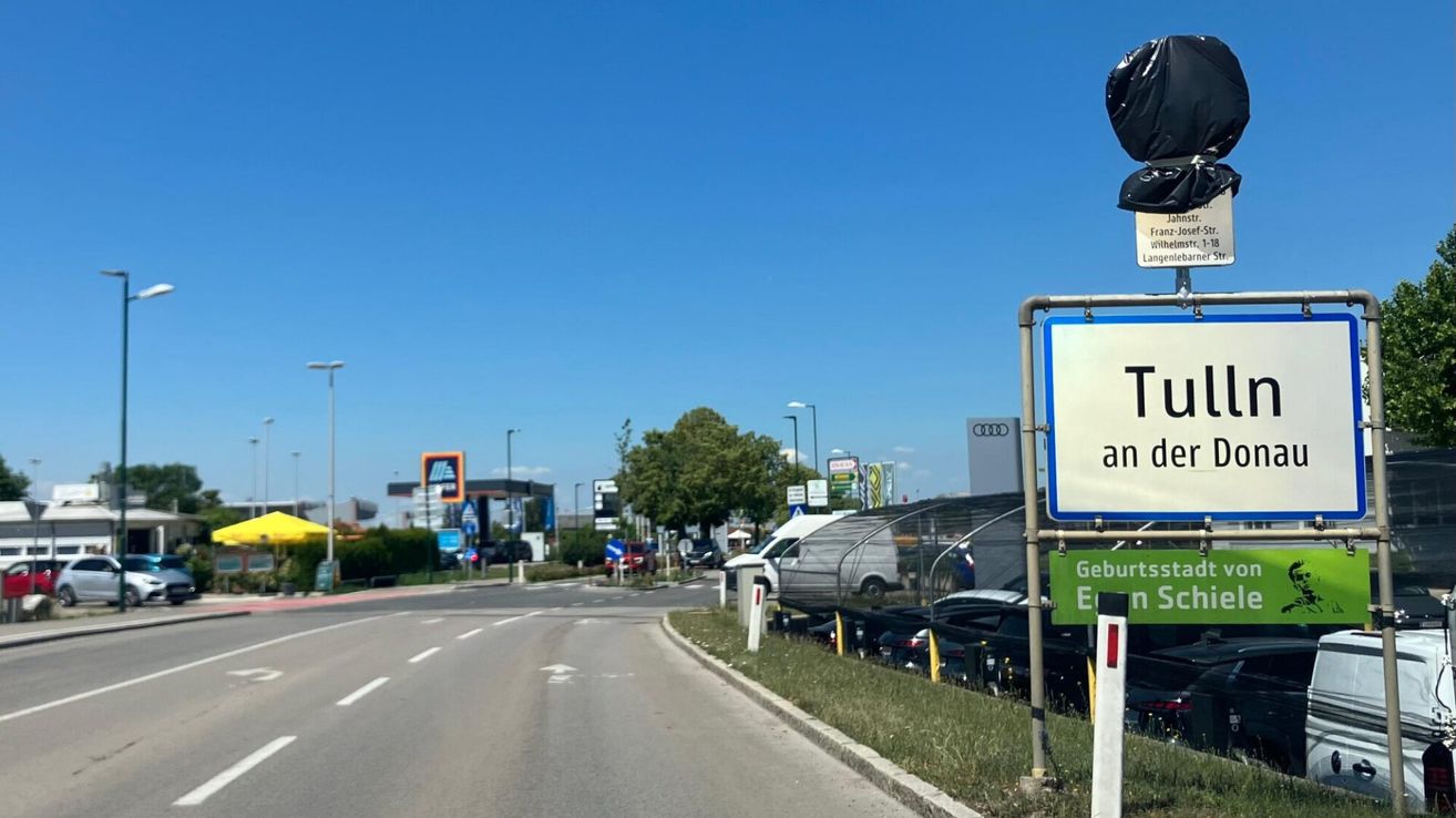 Eine Straße mit geparkten Autos in der Nähe einer Tankstelle. Ein Schild steht 'an' und ein grünes Schild steht 'Gebur E'.