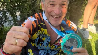 Ein Mann, der auf dem Gras sitzt, zeigt eine Medaille und einen Daumen nach oben. Er trägt ein farbenfrohes Fahrradtrikot, eine Mütze und hat einen Rucksack hinter sich.