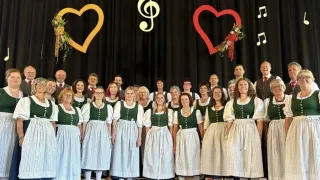 Ein Chor von Frauen und Männern in traditioneller grüner und weißer Kleidung, möglicherweise deutsch, posiert für ein Foto auf einer Bühne mit schwarzem Hintergrund. Musiknoten und Herzen sind auf dem Hintergrund zu sehen.