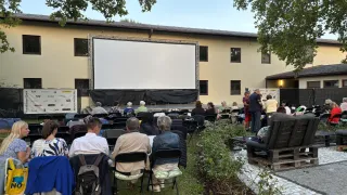 Menschen sitzen auf Stühlen in einem Außenbereich, wahrscheinlich um einen Film auf einer großen Leinwand zu sehen, die an einem Gebäude befestigt ist. Einige stehen in der Nähe der Leinwand. Die Umgebung ist von Bäumen umgeben.