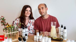 Ein Mann und eine Frau sitzen nebeneinander an einem Tisch. Sie sind umgeben von Flaschen, einer Vase mit Blumen und einem Krug Sahne.