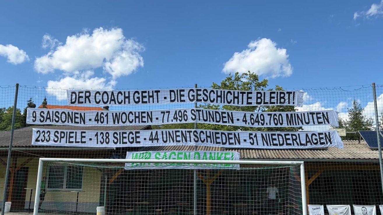 Ein Banner mit deutschem Text hängt am Himmel und zeigt Statistiken. Der Text lautet: Der Trainer geht, aber die Geschichte bleibt. 461 Wochen, 77.496 Stunden, 4.649.760 Minuten. 138 Siege, 44 Unentschieden, 51 Niederlagen. Mit Dank, Danke!
