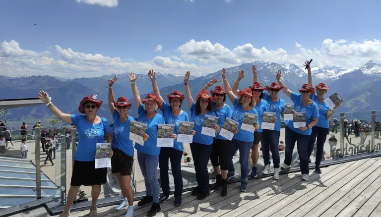 Eine Gruppe von Frauen in blauen T-Shirts und roten Hüten lächelt und posiert für ein Foto auf einer Holzterrasse mit Bergen im Hintergrund.