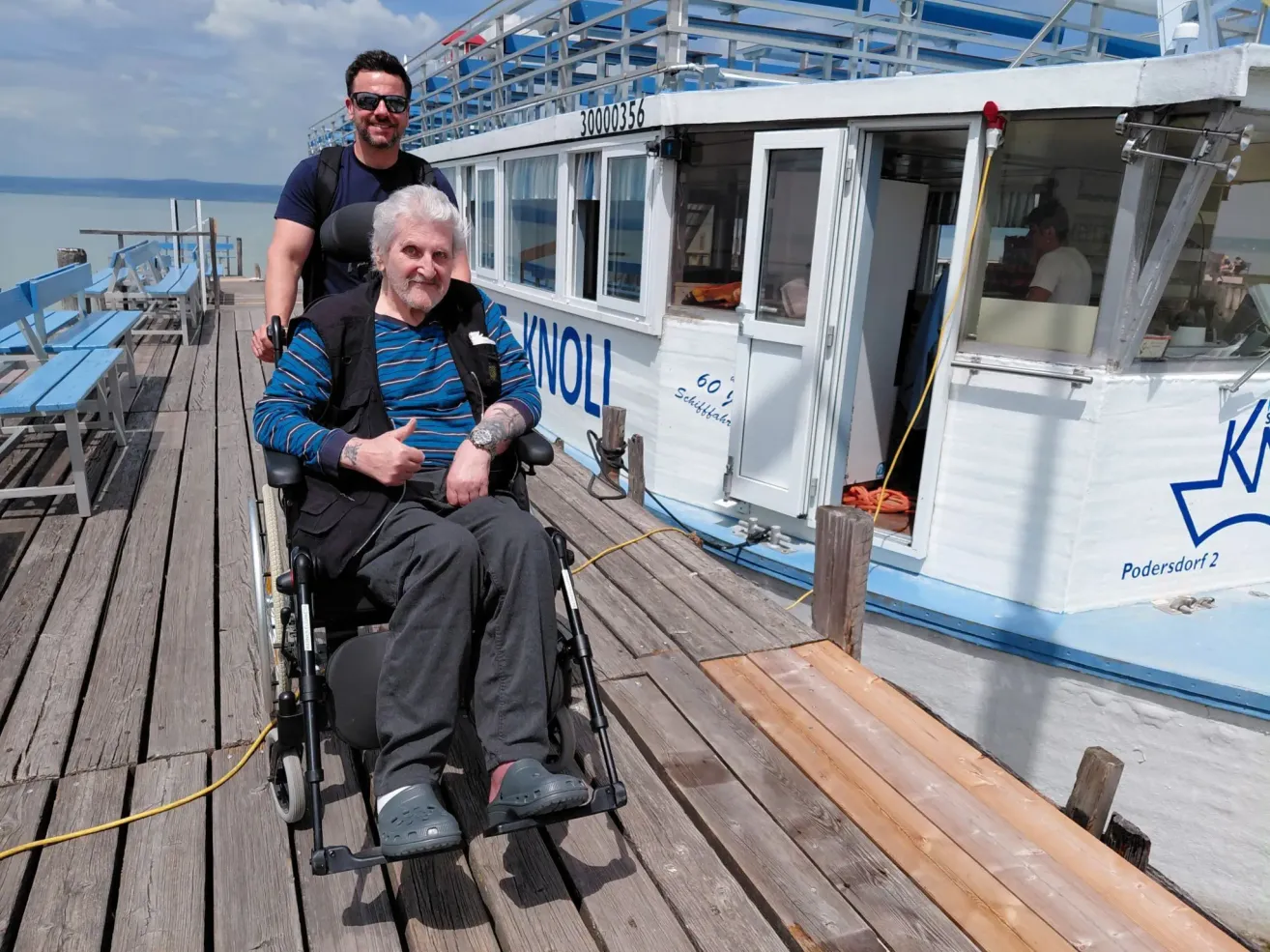 Ein älterer Mann im Rollstuhl gibt auf einem Dock mit einem Boot namens 'KNOLL' im Hintergrund einen Daumen hoch. Ein Mann steht hinter ihm und trägt eine Sonnenbrille.