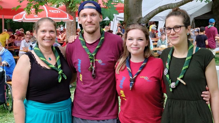 Drei Personen in einem Park mit grünen Lanyards. Der Mann in der Mitte trägt ein weinrotes Shirt. Hinter ihnen sitzen Leute an Tischen unter Schirmen.