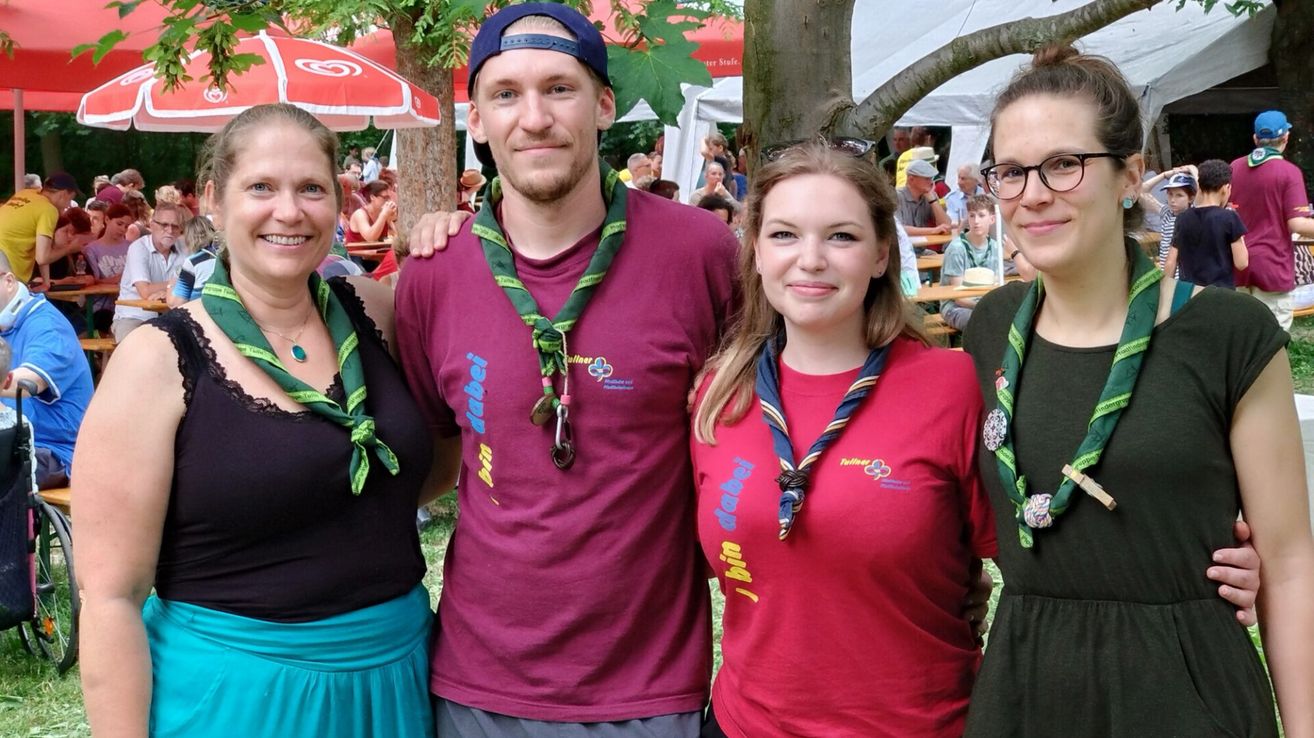 Drei Personen in einem Park mit grünen Lanyards. Der Mann in der Mitte trägt ein weinrotes Shirt. Hinter ihnen sitzen Leute an Tischen unter Schirmen.
