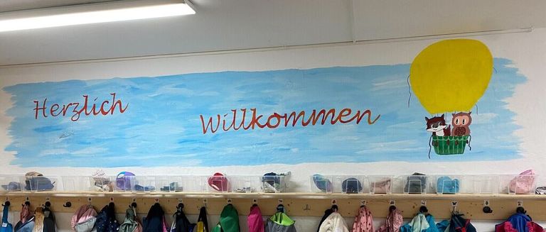 Ein Klassenzimmer mit einem Wandbild, das 'Willkommen' mit einem Heißluftballon und zwei Cartoon-Tieren sagt. Unten steht das Wandbild, eine Reihe von Jacken und Rucksäcken.