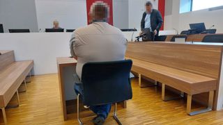Ein Mann sitzt mit verschwommenem Gesicht in einem Gerichtssaal. Ein anderer Mann steht vor einem Schreibtisch mit Monitor. Beide Personen haben verschwommene Gesichter.