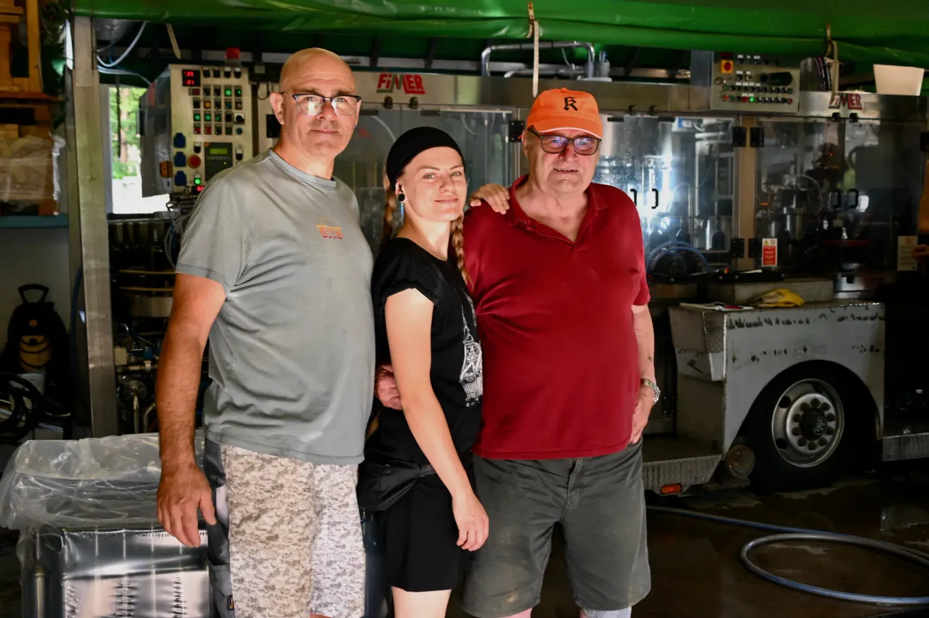 Drei Personen stehen in einer Fabrik zusammen. Ein Mann mit Brille und einem grauen T-Shirt, eine Frau mit einem schwarzen Kleid und ein rotkappener Mann in einem roten Hemd. Sie alle lächeln und scheinen für ein Foto zu posieren. Hinter ihnen befinden sich einige Maschinen.