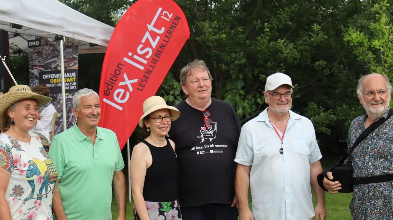 Vier Personen posieren für ein Foto vor einem roten Banner, wobei einer der Männer eine Mütze und eine Brille trägt, ein anderer ein weißes Hemd und eine Brille und eine Frau in der Mitte.