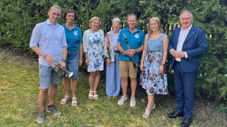 Eine Gruppe von Menschen steht lächelnd in einem Garten, ein Mann hält eine Trompete und ein anderer eine Klarinette. Sie tragen leichte Kleidung, was auf ein zwangloses Treffen hindeutet.