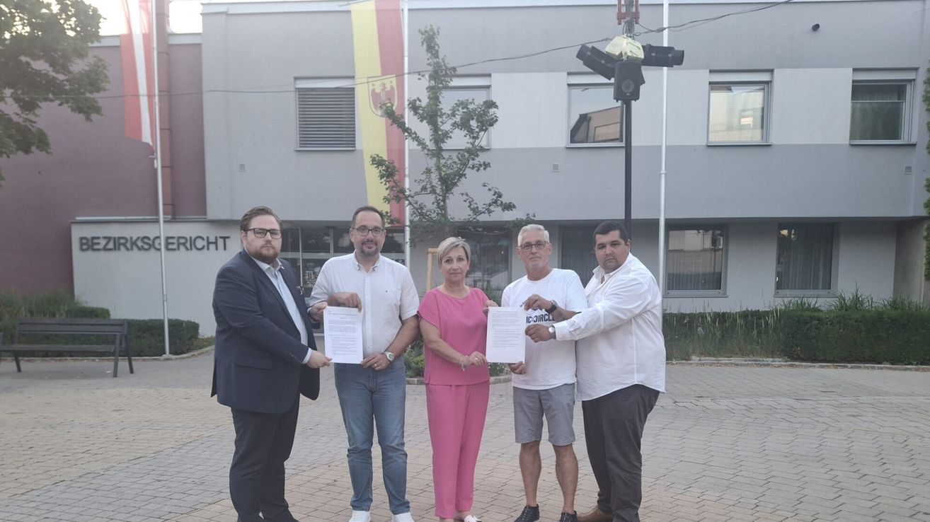 Fünf Personen stehen vor einem Gebäude, halten Papiere in den Händen und lächeln. Der Mann auf der linken Seite trägt einen Anzug, während die anderen Freizeitkleidung tragen. Eine Flagge ist auf einem Pfosten hinter ihnen gehisst.