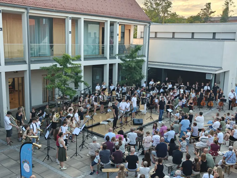Ein Outdoor-Auftritt eines großen Orchesters, mit Musikern, die verschiedene Instrumente vor einem Publikum spielen, das auf Bänken und Stühlen sitzt. Der Hintergrund zeigt ein modernes Gebäude mit Glasfenstern und Balkonen.