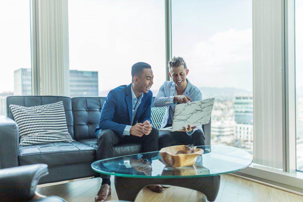 Two men in formal wear sit on a couch, smiling. One holds a laptop while the other points. They sit in a room with a glass table and large windows showing a city view.