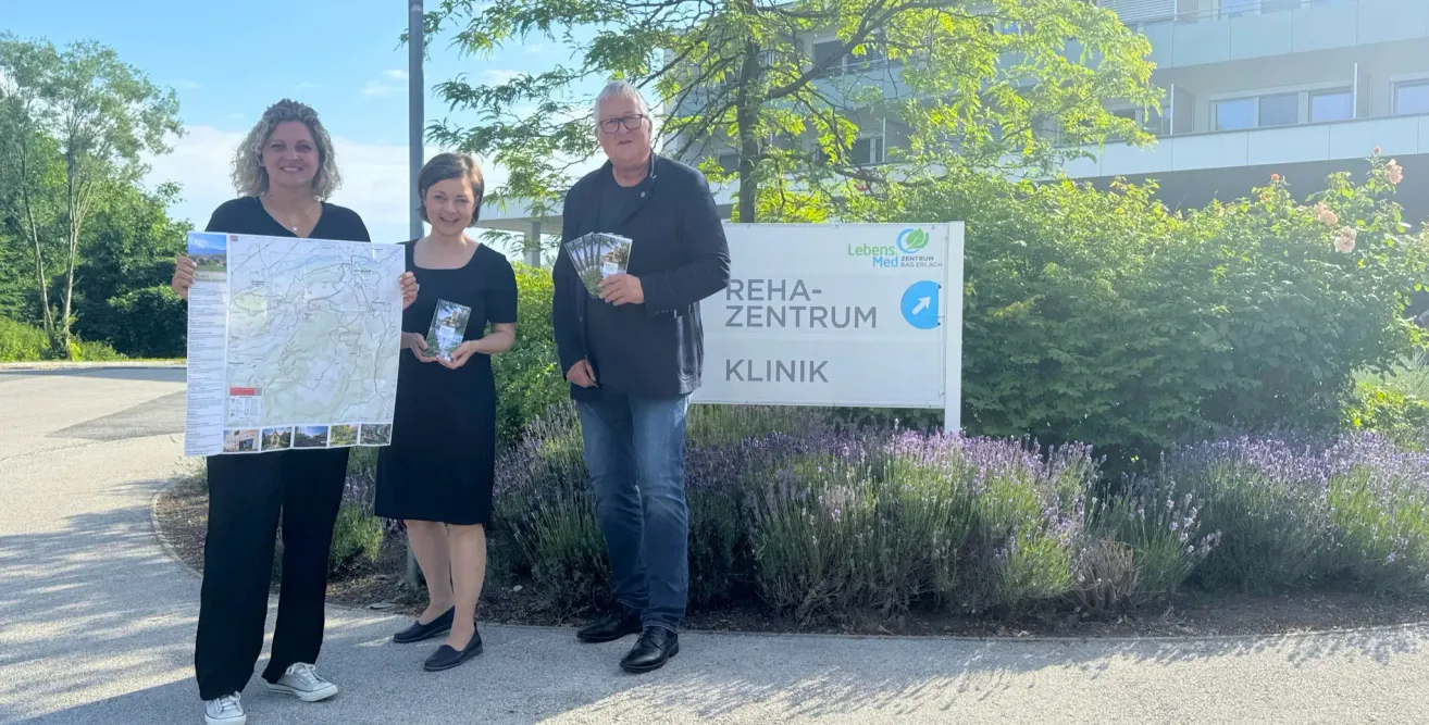 Drei Personen stehen vor einem Schild für das Rehab-Zentrum Klinik. Sie halten Broschüren und eine Karte. Bäume und Blumen sind hinter ihnen.