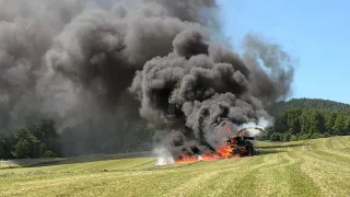 Ein großes Fahrzeug brennt auf einer Grasfläche, wobei dunkler Rauch in den Himmel aufsteigt. Bäume und eine Bergkette sind im Hintergrund zu sehen.