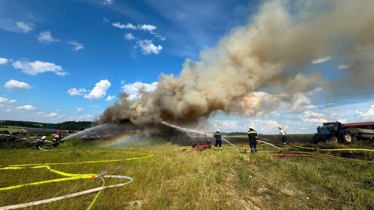 Firefighters extinguish a large fire on a field with a rainbow in the sky, surrounded by smoke and hoses.